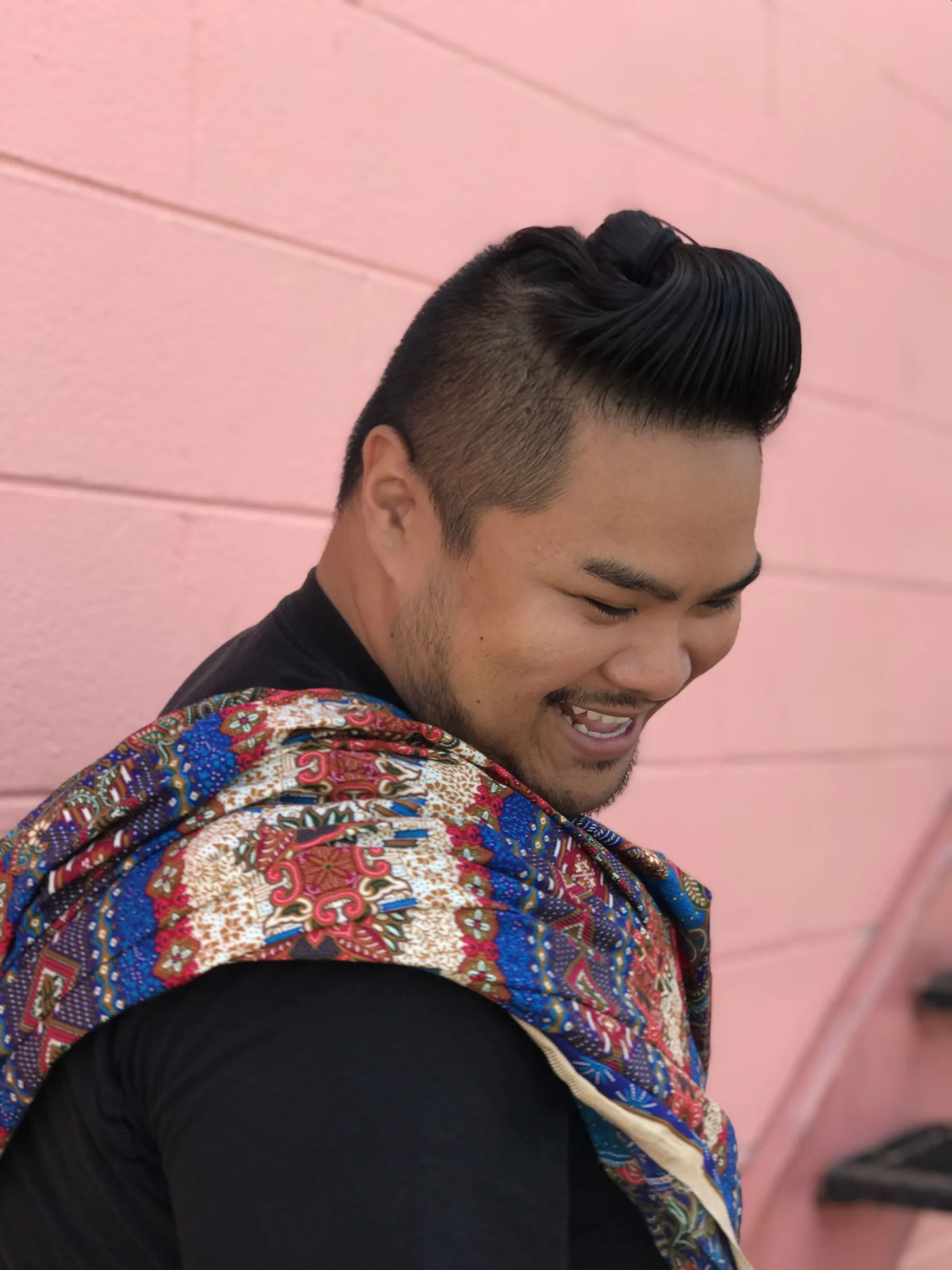 RV Mendoza smiles while looking down in front of a pink wall, wearing a colorful patterned scarf over a black shirt.